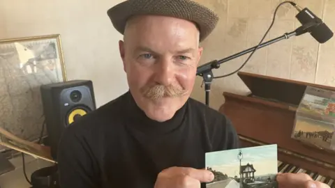 Composer Stuart Wood holding an old postcard from Cleethorpes