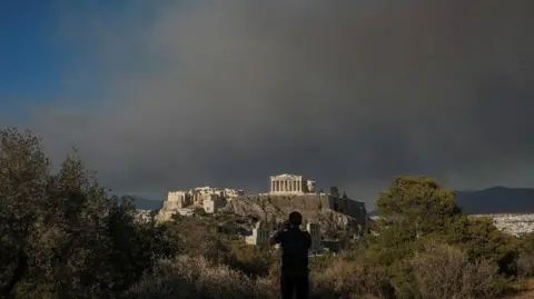 Reuters Een man fotografeert de Parthenon-tempel op de Akropolis-heuvel terwijl er rook te zien is van een bosbrand in het dorp Varnava in Athene, Griekenland, op 11 augustus 2024.