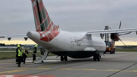 Fubar News A turboprop aircraft with a red and green patterned tail parked on an airport taxiway, surrounded by ground crew in high-visibility jackets, under an overcast sky