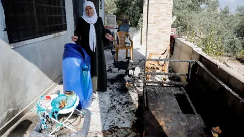 Reuters A Palestinian woman gestures towards furniture and a baby walker that were burned in an attack by Israeli settlers in the village of Jit in the occupied West Bank (August 16, 2024)