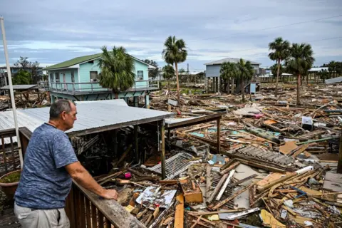 CHANDAN KHANNA/AFP Seorang pria memeriksa kerusakan rumahnya pasca Badai Helene. Gambar tersebut menunjukkan puing-puing dari rumah-rumah sejauh mata memandang.