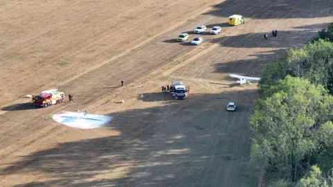 The scene of an aircraft crash at Adversane, south of Billingshurst, on Monday 10 August. A fire engine, ambulance and other emergency service vehicles are visible from a bird's eye view. The white aircraft can be seen in the image.