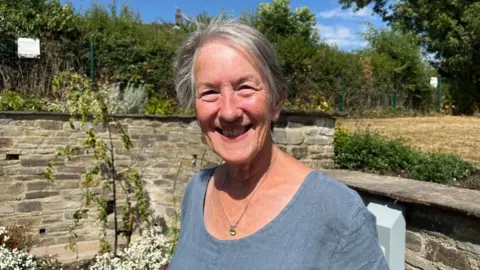 Ellen Knight/BBC Martin photographed looking into the camera and smiling. She is wearing a light blue linen top, and is stood in front of a low stone wall with plants and flowers growing up it. The sky is blue and there is strong sunshine. 