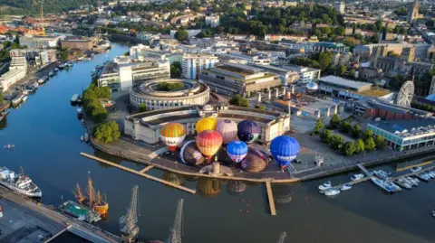 PA Media A drone image of Bristol Harbourside of hot air balloons on land. The River Avon which passes through harbourside is shown. 