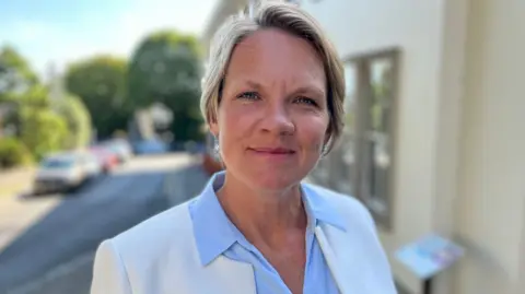 Deputy Sasha Kazantseva-Miller - A woman with short cropped blonde hair, wearing a white suit jacket with no collar and a blue shirt. 