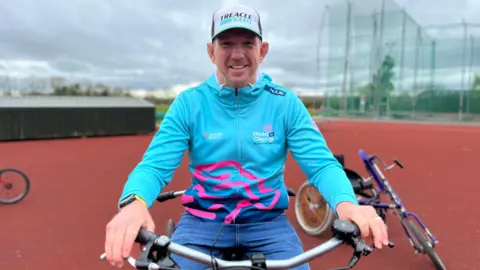 A man in his mid 40s sits on an adapted bike at an athletics track. The surface is red and other bikes can be seen around him, with a throwing cage behind. He is wearing a white cap, a bright blue and pink jacket, and jeans. He is smiling.