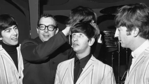 Getty Images Eric Morecambe is dressed in a Beatles-style coat and holding a Beatles-style wig above the head of Ringo Starr as George Harrison and John Lennon look on from either side.