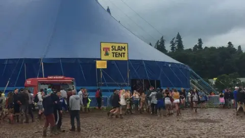 People walk across mud and enter a large blue tent at a music festival