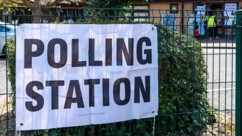 Getty Images Picture of outside a polling station - a sign is in the foreground and in the background you can see the polling station open, with the rules in the window for voting. 
