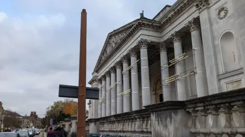 Alex Harris/BBC The front of the Fitzwilliam Museum has ornate columns outside. On the path outside is a post which is missing the lamp on top. There are people walking on the pavement and cars driving down the road