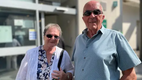 Barbara Havard (left) and Mike Quaggan are an older couple, standing together on a sunny day in front of the doors to a bank. They are smiling wearing sunglasses.