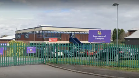 Road leading up to green gates that are closed around a school. The school is built from red bricks and has a purple sign that reads "Lawnswood Campus" to the left.