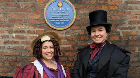 Two women, who are dressed up as Ann Walker and Anne Lister, are smiling to the camera in front of a rainbow plaque honoring Anne Lister. 
