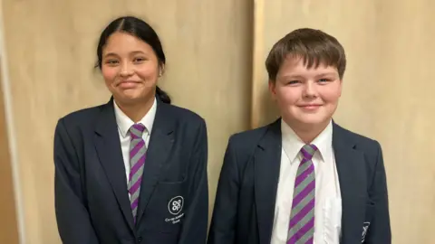Two school children in a navy blue blazer, white shirt with a purple and grey striped tie on.