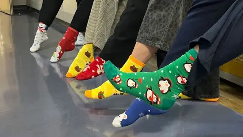 Katherine Ganczakowski/BBC A close up of seven left legs, all wearing brightly coloured Christmas socks and pointing their toes. 