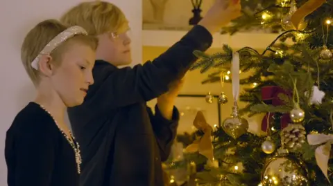 Blatella Films / Gold Geese A side-on candid photo of a boy and a girl putting decorations on a Christmas tree, both are dressed smartly and focused on their task