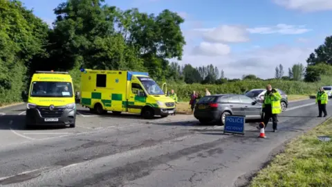 The scene of a car crash with a 'police accident' sign and emergency service vehicles and workers