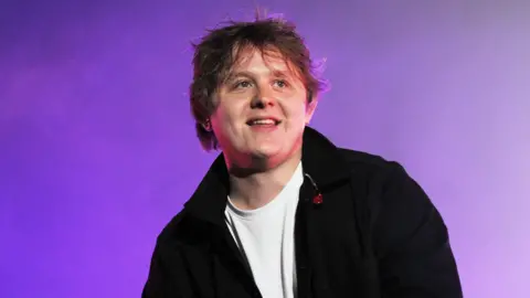 Lewis Capaldi performs onstage - he is wearing a white shirt and black jacket, is holding a microphone and smiling