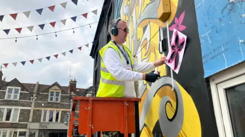 The graffiti artist Inkie stands on a cherry picker and holds a stencil of a flower against a wall that he is about to spray with pink paint. There is already a black and yellow design on the wall. There are shops in the background and lines of bunting behind him.