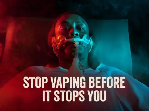Gov.sg / Stop Vaping An ominous looking public health advertisement showing a man on a hospital bed with an oxygen mask surrounded by smoke and red and blue lighting, with the tagline "Stop vaping before it stops you".