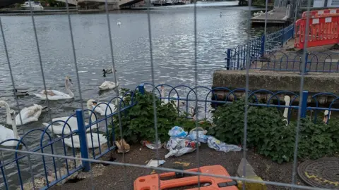 CWRS A view of Caversham Bridge, with swans in the water. A metal barrier and rubbish is by the river edge.