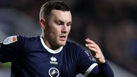 Salford City striker Macaulay Langstaff pictured in his dark blue Millwall strip, arms pumping as he chases the ball.