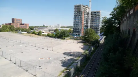 A large empty concrete space with fencing on it. Construction work is ongoing on a block of flats in the background.