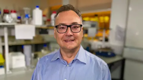 Martin Giles/BBC Professor George Malliaras in a lab. He has short grey hair, dark framed glasses and is wearing a lilac shirt. He is smiling into the camera.