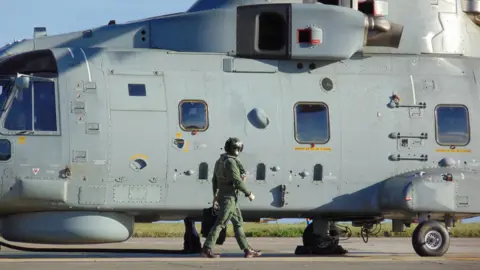 RNAS Culdrose A pilot wearing a dark green jumpsuit and black helmet and holding a case is walking along the length of a grey Merlin helicopter while it is stationary on the runway.