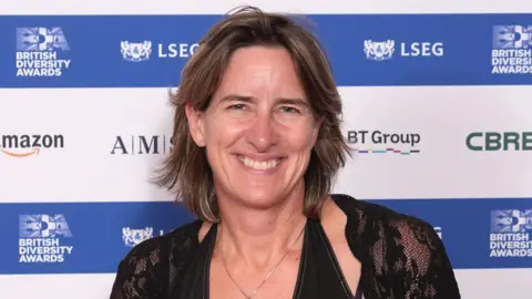 PA Media A smiling Dame Katherine Grainger looks directly at the camera as she is photographed in front of a step and repeat banner which has the logos of the brands Amazon, British Diversity Awards and BT Group displayed. She is wearing a black lace shawl over a black dress, with a silver necklace. 