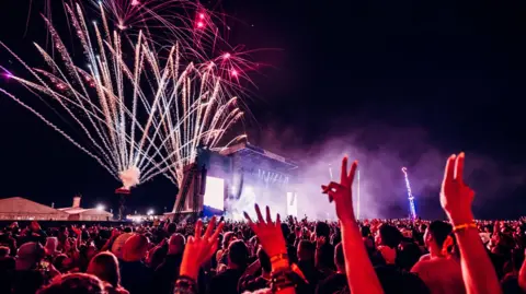 A photo taken from in the crowd of as a night-time act is underway. There are orange, pink and white fireworks going off behind a stage which has dramatic white show lighting. In the foreground are people in the crowd raising their hands. 