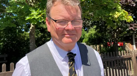 A man with glasses and grey hair looks to the camera and smiles. He is wearing a white shirt, grey waistcoat and dark tie with yellow stripes. He is beside a fence and tree and is in the sunshine.
