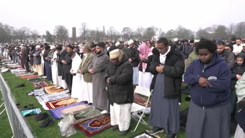 Rows of men lined straight in a park stood on prayer mats. 