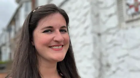 Headshot of Eleanor Bryan with long brown hair smiling with teeth into the camera in front of the house