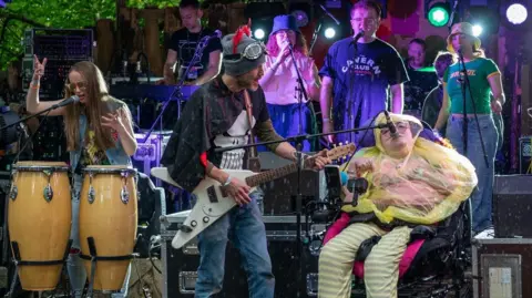 Barry Fahey The band are performing on stage at a festival. The young woman in the right of the picture is singing into a microphone. She is wearing a yellow rain poncho and is a wheelchair user. Beside her is a man playing an electric guitar and to his left is another young woman who is playing Conga drums. Behind them other young people are singing into microphones and playing instruments. 