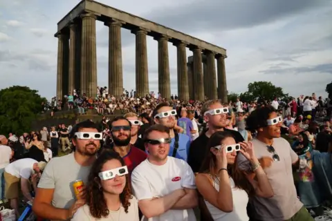 PA Media Crowds of mixed genders and ages stand on a grassy hilltop with the columns of the National Monument visible in the background. A group of friends at the front can be seen wearing eclipse glasses.