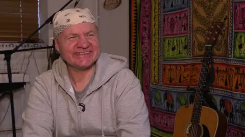 Shaun Hayward, who is wearing a grey zip-up hoody and a black and white bandana, smiles at the camera. In the background an acoustic guitar leans against a large, colourful rug on the wall