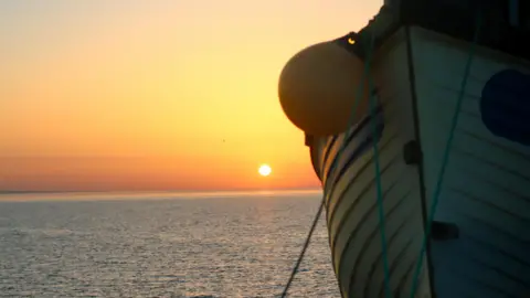 Weather Watchers/RECROSS The hull of a boat, tied up on land. A calm sea can be seen behind it, with the sun having just risen above the horizon. The sky is orange and yellow.