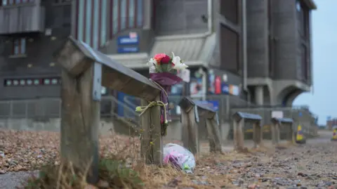 Eddie Mitchell A bouquet of flowers is tied to a wooden beach barrier on a pebble shoreline near Shoreham RNLI. Buildings and seafront structures can be seen in the background.