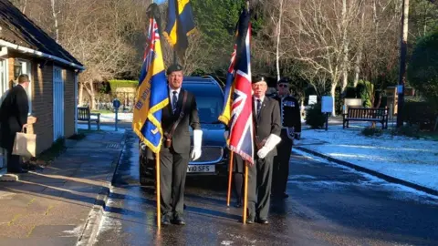 Jacky Carter Four people in suits and berets stood in front of a hearse. they are carrying British flags.