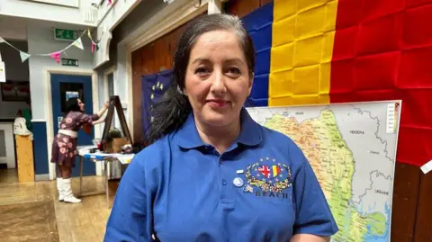 Andreea Salvage, the organiser of the Sunday school, is looking at the camera. She has black hair tied in a ponytail and is wearing a blue t-shirt with a heart on the front. One half of the heart is the UK flag. The other half is the Romanian flag.