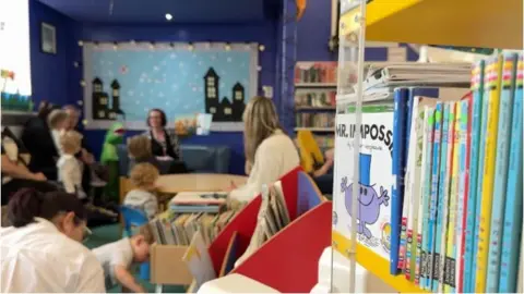 A story time session at the family library, you can see a bookshelf in the foreground and children and parents in the background listening to a woman read a story. She holds a book up.