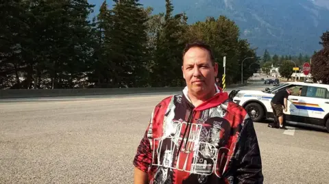 Karl Hasketh A man stands on the roadside wearing a red and black graphic hoodie with trees, mountains, and a police car behind him.