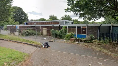 A derelict single-storey school building with metal fencing surrounding it and litter on the ground