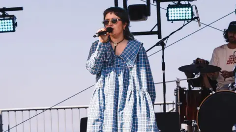 Essy Sparrow in a blue and white checked dress is standing on a stage while singing into a microphone as a drummer drums behind her.