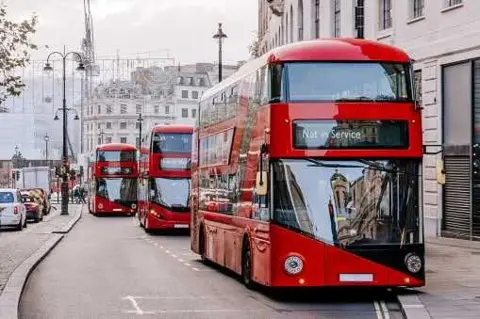 Three red Transport for London buses on a street in central London. The bus at the front is out of service and parked in a bus lane. In the background there are lampposts, buildings and parked cars on the opposite side of the street. 