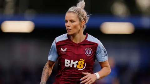 A woman playing football while wearing a claret and blue Aston Villa shirt. Her blonde hair is tied back and she is looking to the right with her left arm partially raised. She has tattoos on her right arm.