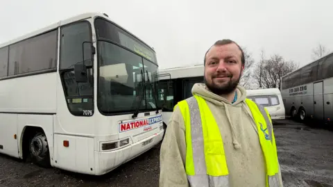 BBC/Jim Scott Callum Bowes, wearing a green hoodie and a yellow high visibility jacket, smiles. He is standing near a white former National Express coach, which has some dirt marks on the front of it after being transported to Shotton Colliery.