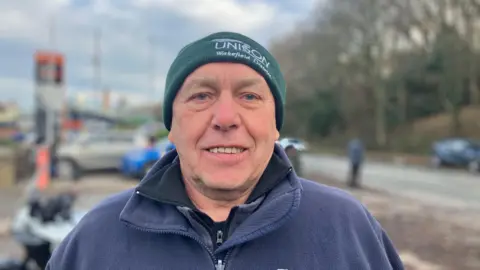Elizabeth Baines/BBC A man with a blue fleece and a wooly hat smiles at the camera. His hat has UNISON written on it.