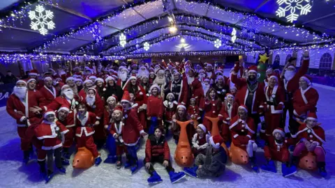 A group photo of people dressed as Santa on an ice rink at Clarks Village in Street. Some of them are seated on the ice and many are standing and smiling at the camera. The picture is taken at night and the rink is lit up by illuminations overhead, some of them shaped like snowflakes